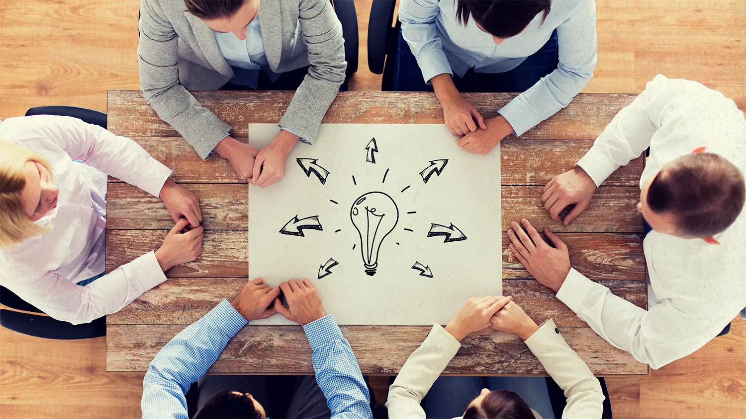 Image of 6 business people sitting around a table with a paper in the center featuring a lightbulb sketch with arrows pointing in different directions representing talking to a business consultant.